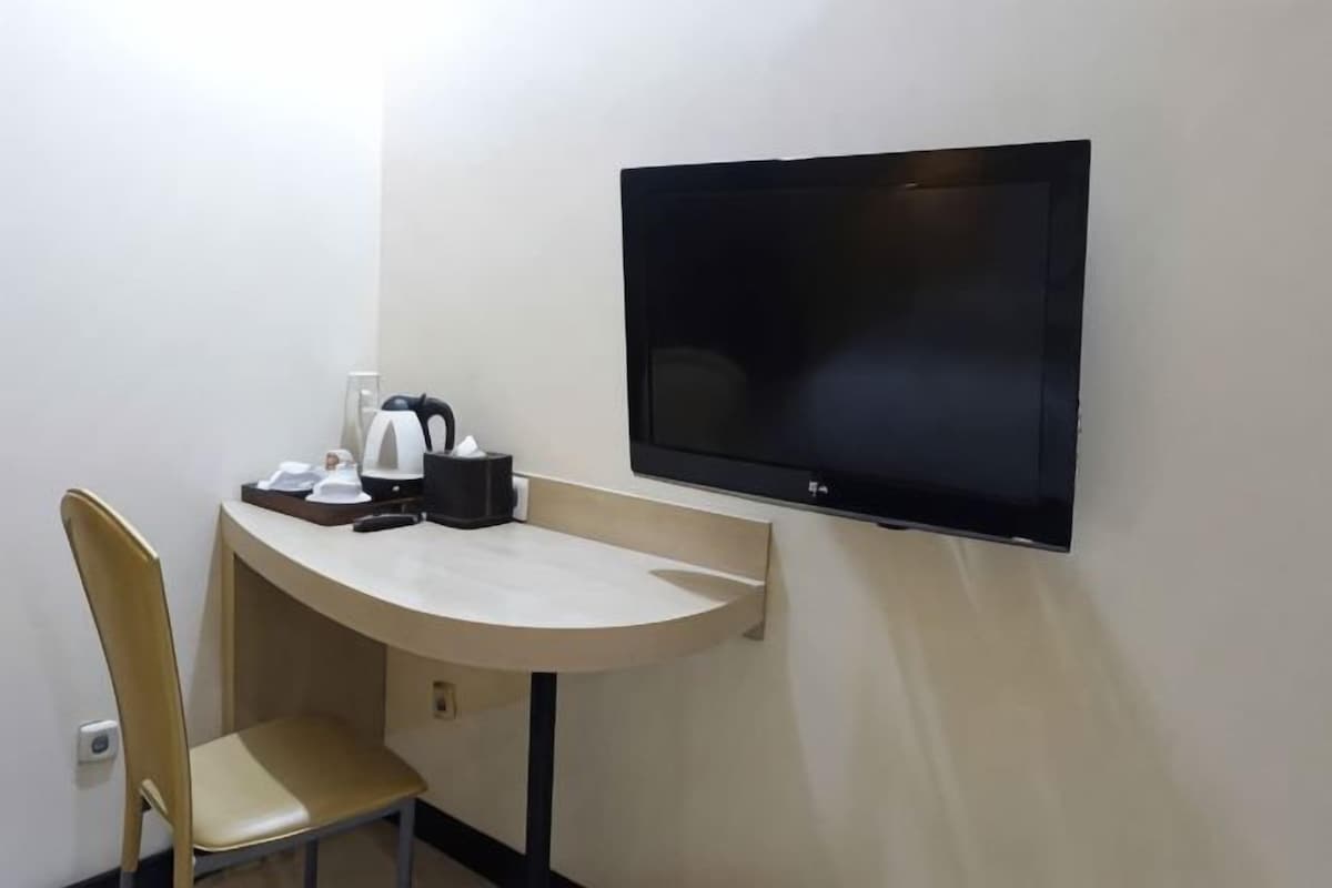 A hotel room with a flat-screen TV mounted on the wall, a small table with a kettle, a cup, and a tray, and a single chair.