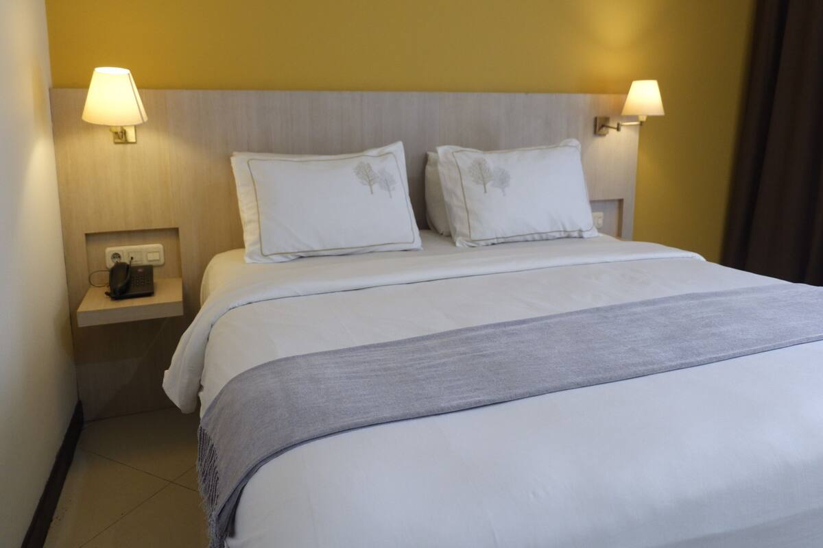 Quarto de hotel com uma cama, duas luminárias de cabeceira, um telefone em uma mesinha de cabeceira e um ponto de luz fixado na parede.