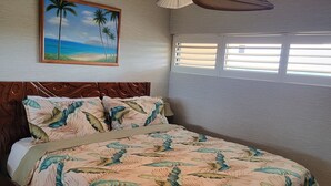 Une chambre à coucher avec un lit, un ventilateur de plafond, une fenêtre avec des stores et un tableau représentant une scène de plage.