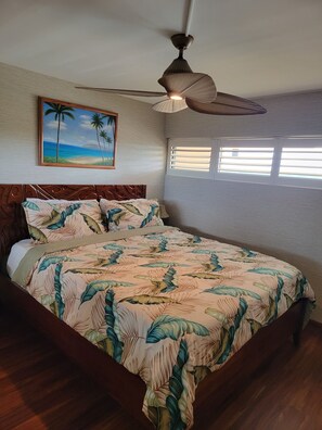 A bedroom with a bed, a ceiling fan, a window with blinds, and a painting of a beach scene.