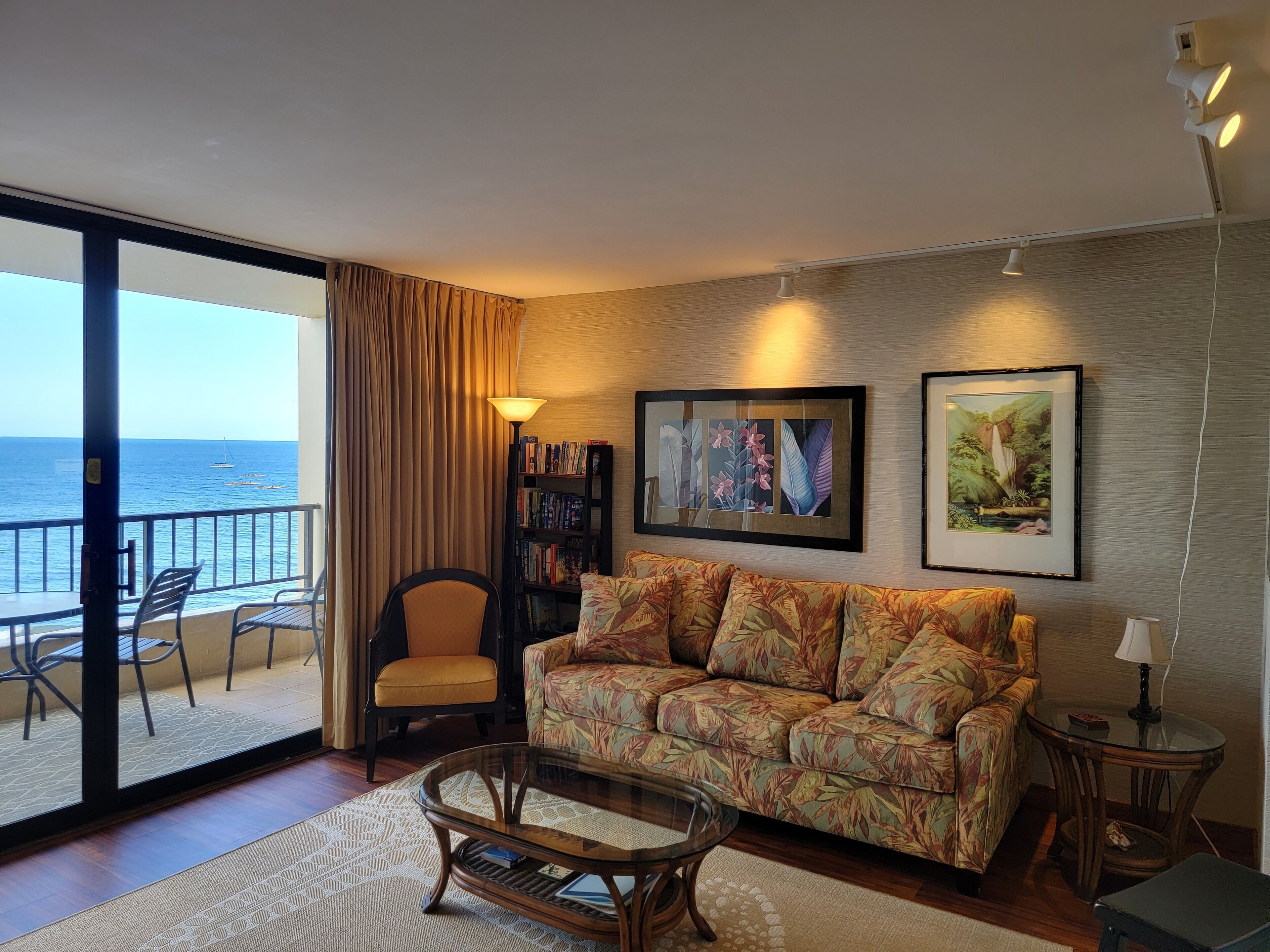A living room with a sofa, a glass coffee table, a bookshelf, and a view of the sea.