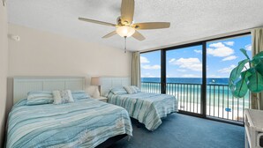 Camera d'albergo con due letti, una ventola a soffitto, un'ampia finestra vista spiaggia e una pianta in vaso.