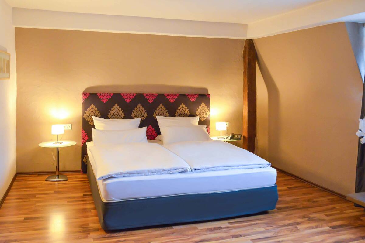 Una habitación de hotel con una cama, lámparas de noche, una mesita de noche y una planta en maceta.