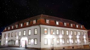 Un grand bâtiment blanc, doté de nombreuses fenêtres et d’un toit recouvert de tuiles rouges, illuminé par des éclairages extérieurs.