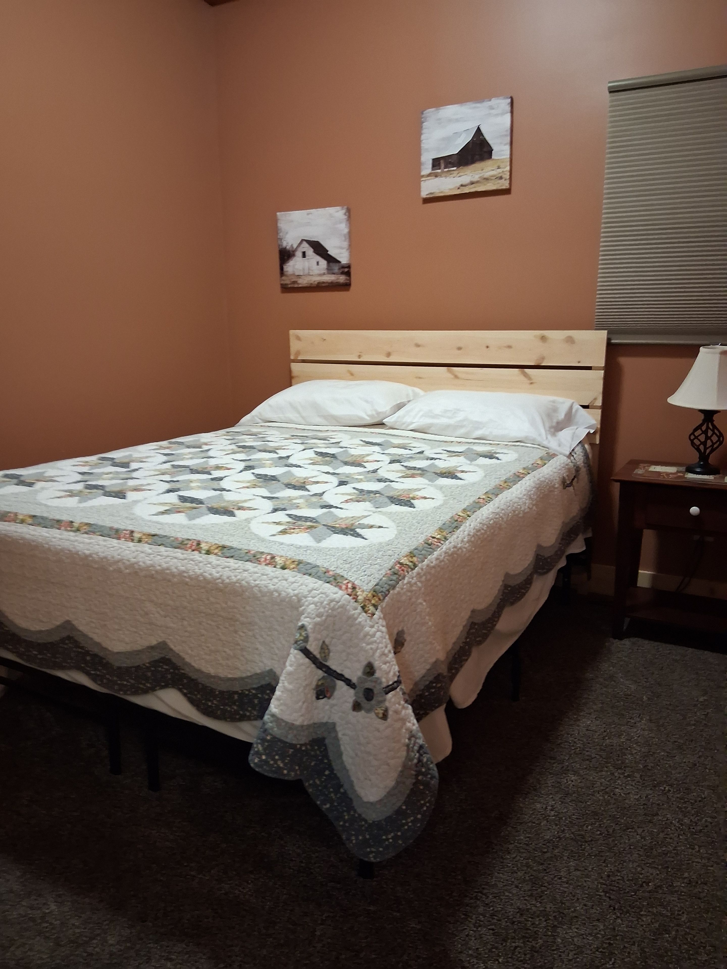 A single bed with a quilt, a wooden headboard, a nightstand with a lamp, and two wall-mounted pictures.