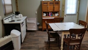 A compact kitchen with a refrigerator, stove, and sink. There is a dining table with chairs and a wooden cabinet with shelves.