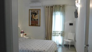 Un dormitorio con cama, mesita de noche, lámpara, silla, ventana con cortinas y un aire acondicionado instalado en la pared.