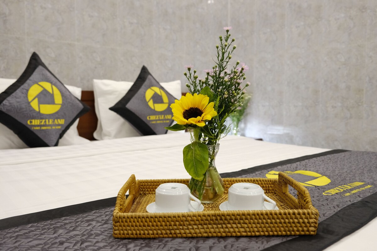 Een hotelkamer met een bed, kussens met een logo, een dienblad met kopjes en een vaas met bloemen.