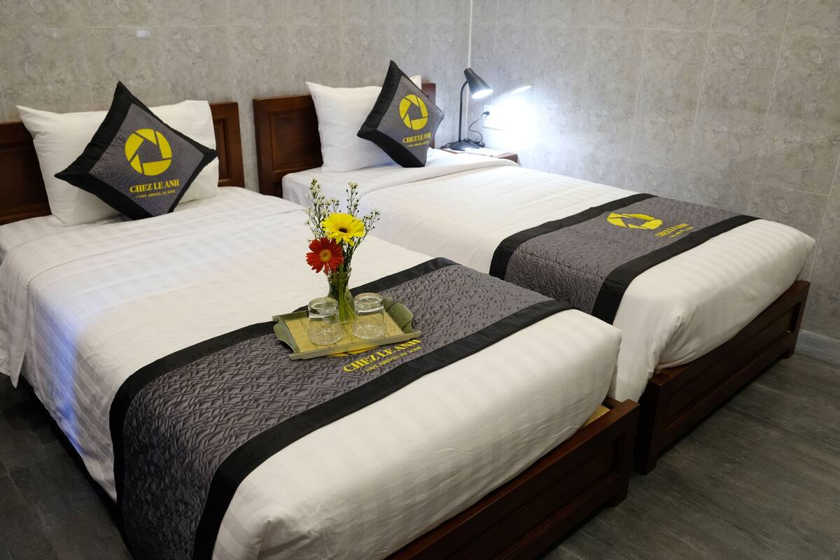 Habitación de hotel con dos camas, cada una con colcha gris y negra, almohadas blancas y un logo en las almohadas. Hay una bandeja con un florero y vasos sobre la cama.