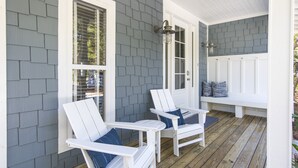 Eine Veranda mit zwei weißen Adirondack-Stühlen, einer Bank und einer Tür mit Fenster.