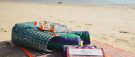 A beach scene with a woven bag, a bottle, and a plaid bag on a blanket.