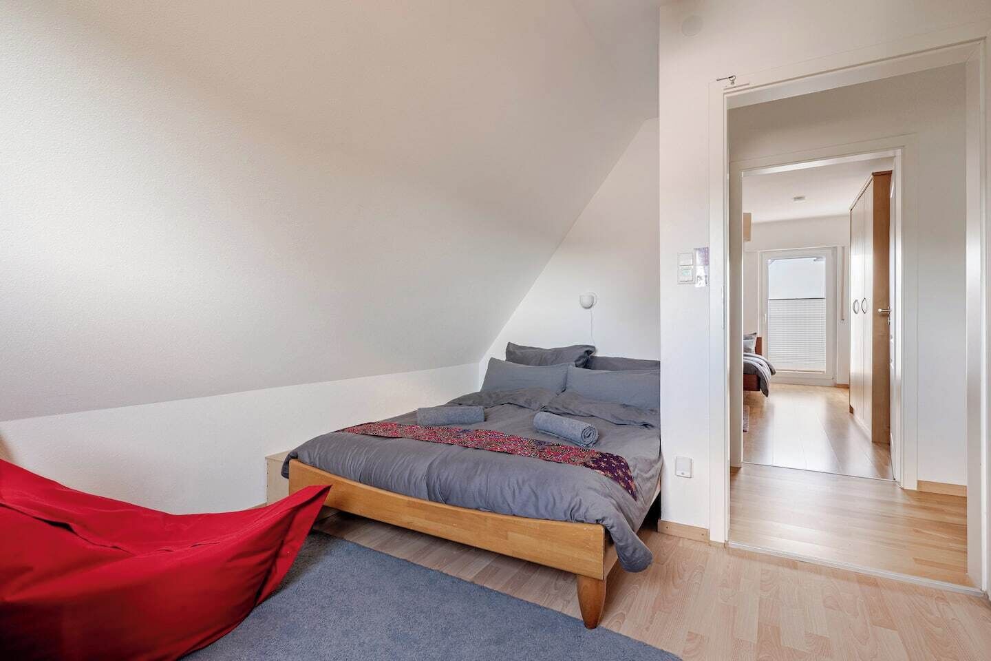 Una camera da letto con soffitto spiovente, un letto con biancheria grigia, una sedia rossa e un pavimento in legno.