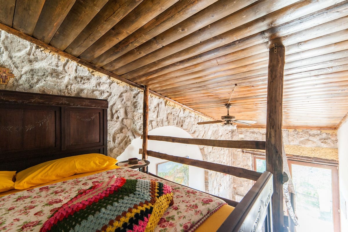 Una habitación rústica con techo de madera, una cama con manta de colores, cabecera de madera y pared de piedra.