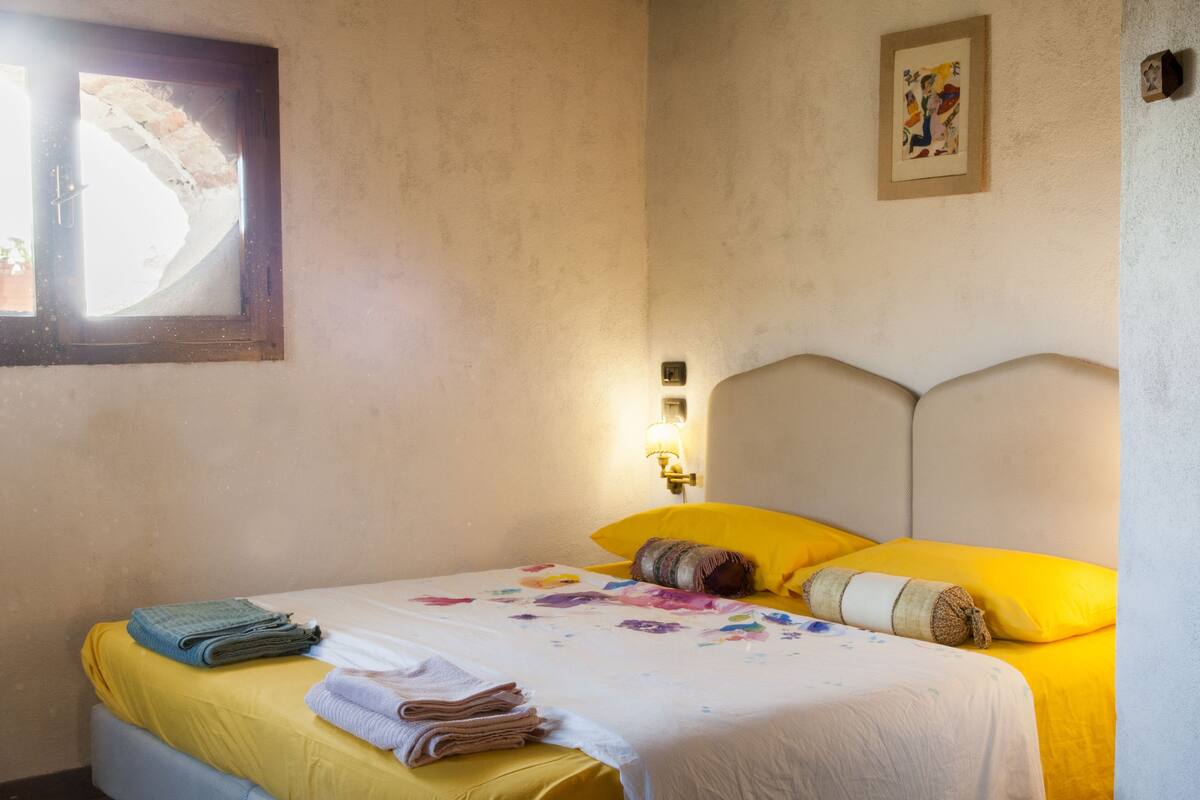 Un dormitorio con cama, almohadas, una mesita de noche, una lámpara y una ventana.