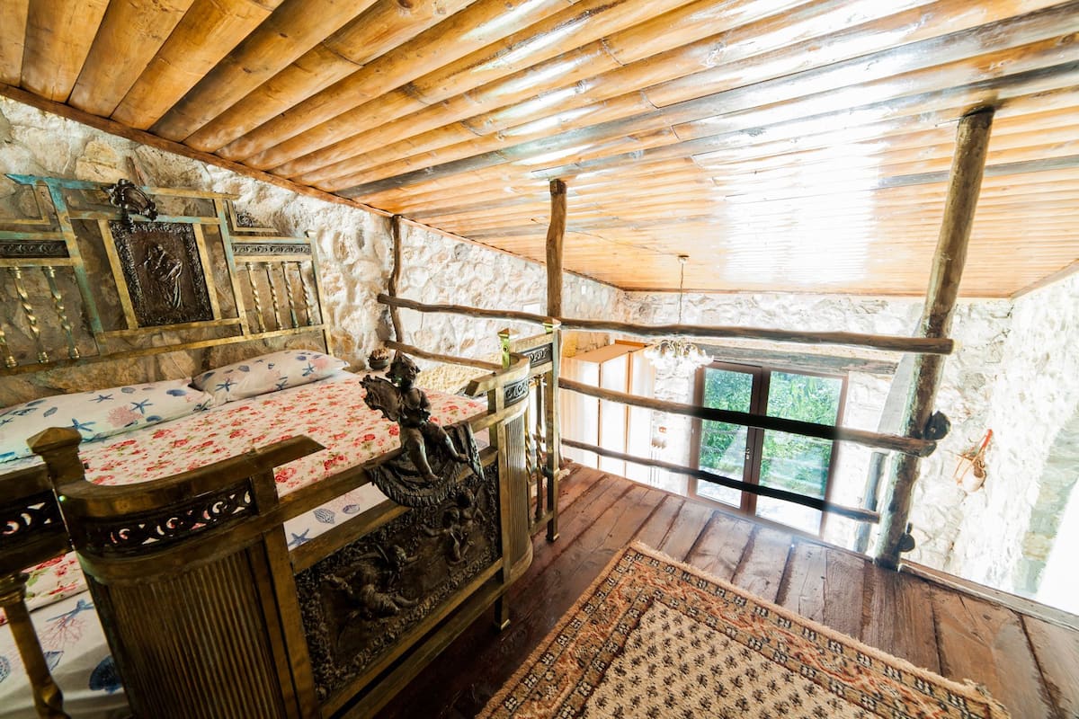 Una habitación rústica con techo de madera, paredes de piedra, una cama con ropa de cama floral y un ventanal grande.
