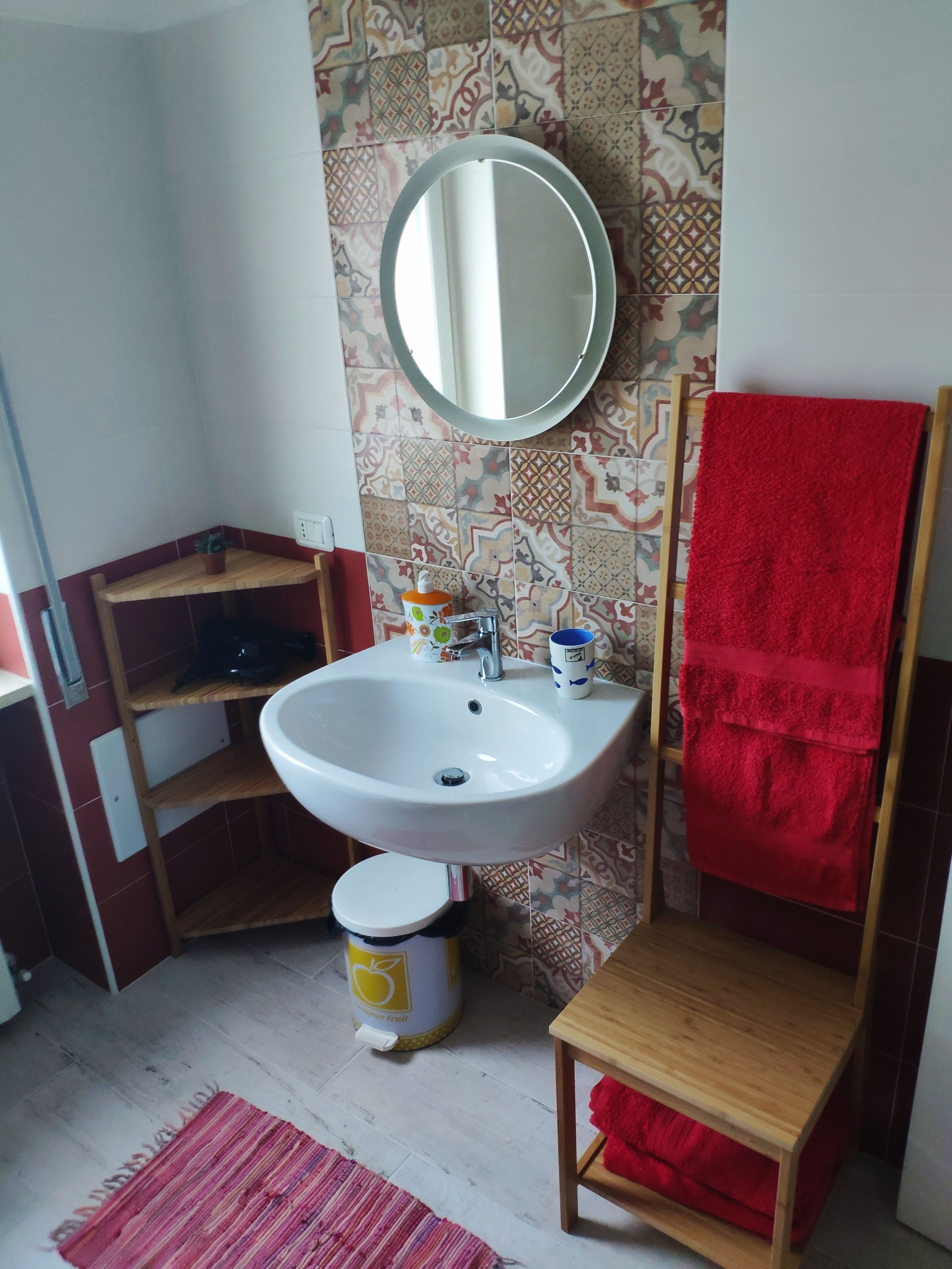 A bathroom with a white sink, a mirror, a red towel, a wooden shelf, and a patterned wall.