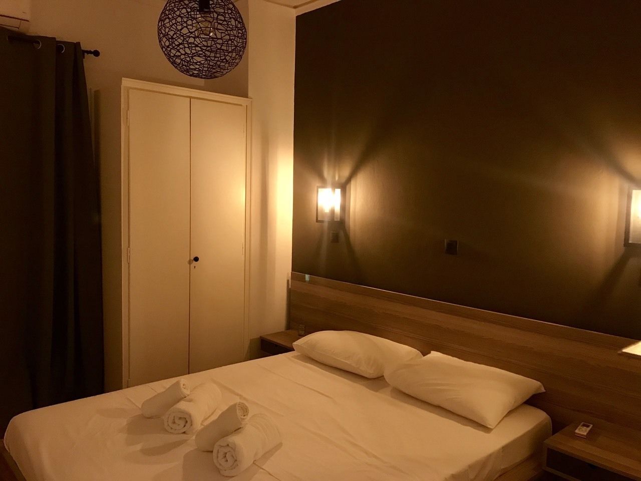 Quarto de hotel com cama bem arrumada, cabeceira de madeira, luminária na parede e porta fechada.