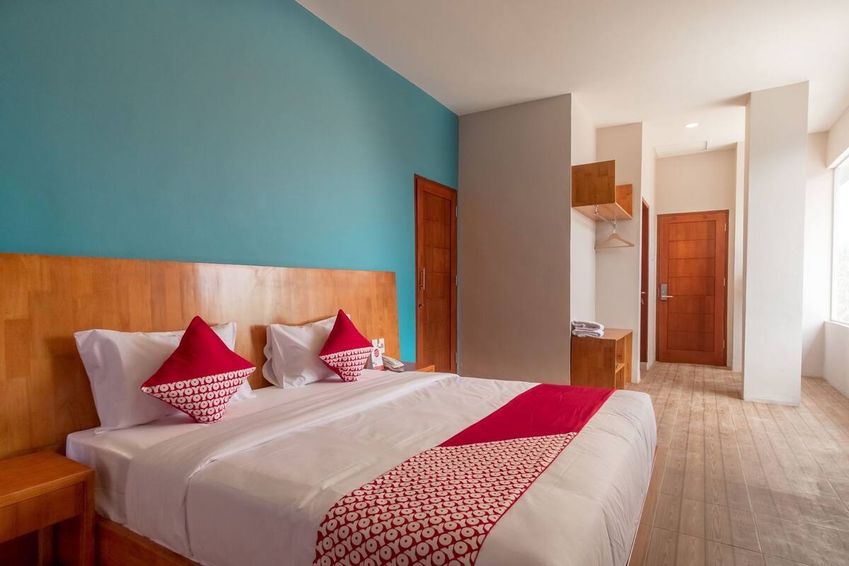 Hotellihuone, jossa on suuri sänky, puinen pääty, punavalkoinen kuviollinen päiväpeite, puinen yöpöytä ja näkymä käytävälle.