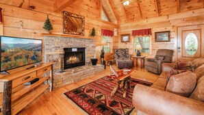 L’intérieur d’un chalet en bois, avec une cheminée en pierre, un téléviseur, un canapé et un ventilateur de plafond.