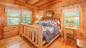 Une chambre à coucher dans une cabane en bois, avec un lit, deux fenêtres avec des rideaux, une table de chevet avec une lampe et un miroir sur le mur.