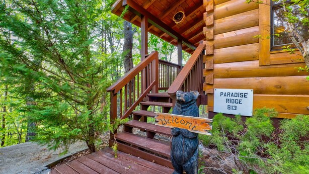 Une cabane en bois avec un escalier en bois et une statue d’ours tenant une pancarte sur laquelle on peut lire : « Bienvenue à Paradise Ridge 813 ».