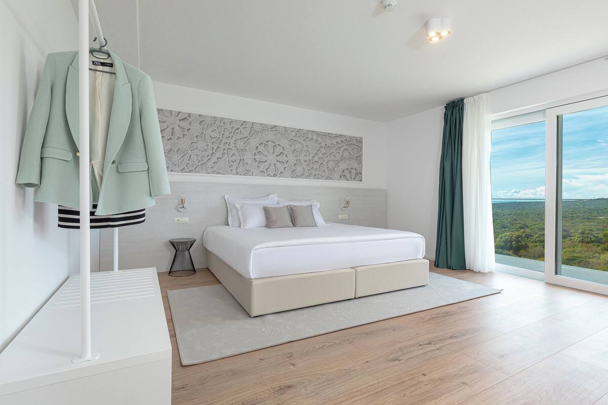 A modern bedroom with a large bed, a hanging coat rack, and a view of the landscape through a large window.