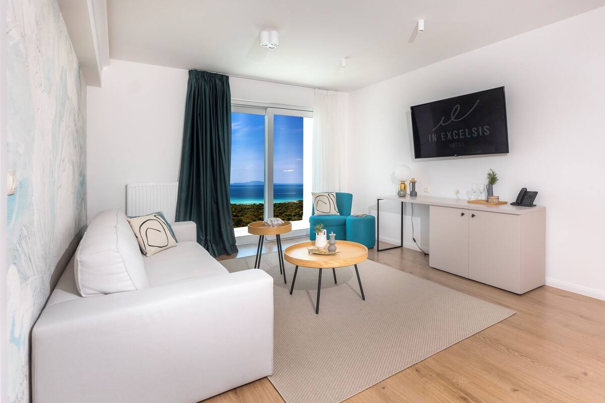 A modern hotel room with a white sofa, a round wooden coffee table, a teal armchair, and a view of the sea through large windows.