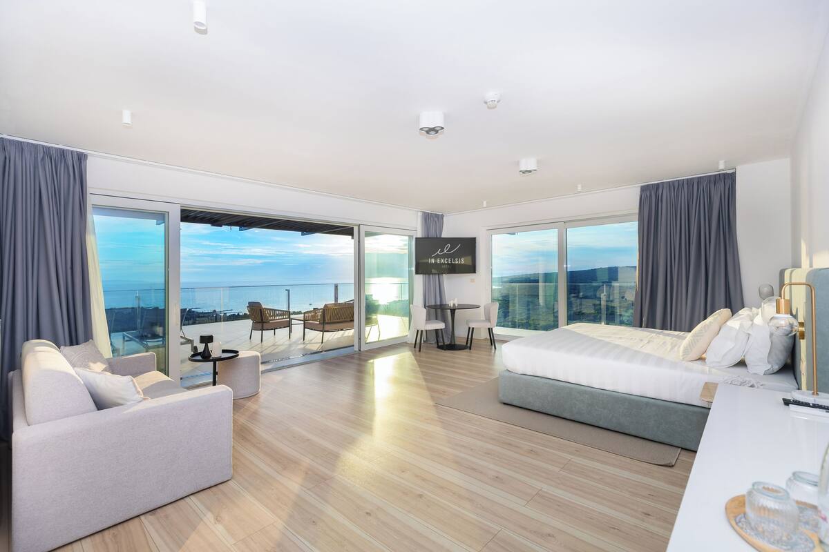 A modern hotel room with a large bed, a sofa, a dining table, and a TV. The room has large windows offering a view of the city.