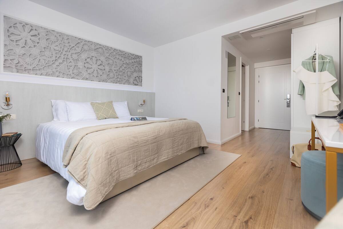 A modern bedroom with a large bed, a decorative wall panel, a bedside table with a lamp, and a wardrobe with a mirror.