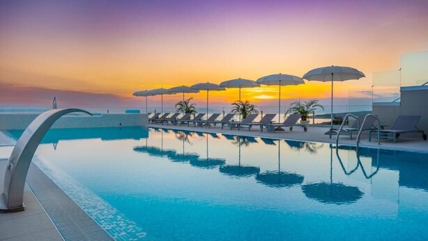 A poolside area with lounge chairs and umbrellas, reflecting a sunset over the water. - Hotel In Excelsis (Novalja)