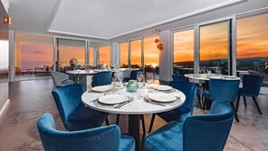 A modern dining area with round tables and blue chairs, large windows offering a sunset view. - Hotel In Excelsis (Novalja)