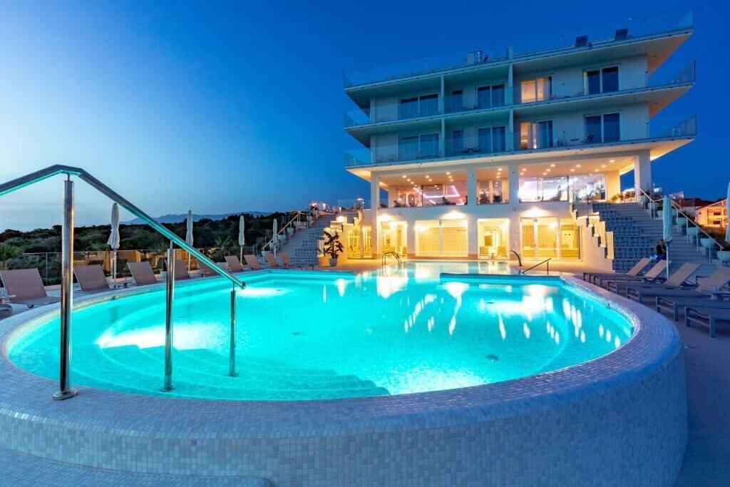 Una piscina con borde de cristal, sillas de descanso y un edificio de varios pisos con balcones al fondo.