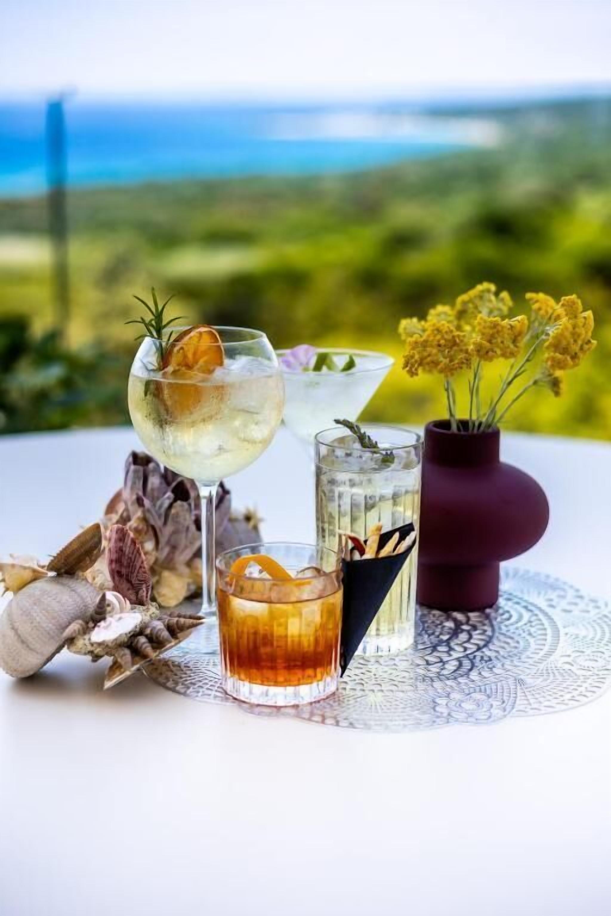 A table with three glasses of beverages, a vase with yellow flowers, and dried herbs.