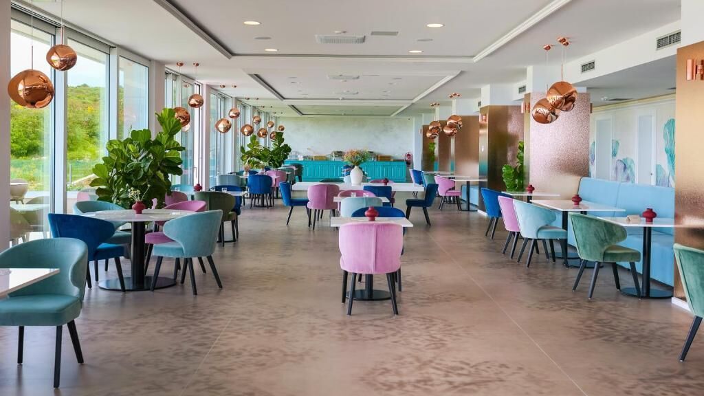 En moderne restaurant med runde borde og farverige stole, store vinduer og dekorative kobberpendler.