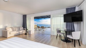 A modern hotel room with a large bed, a sofa, a dining table with chairs, a TV, and a view of the ocean. - Hotel In Excelsis (Novalja)