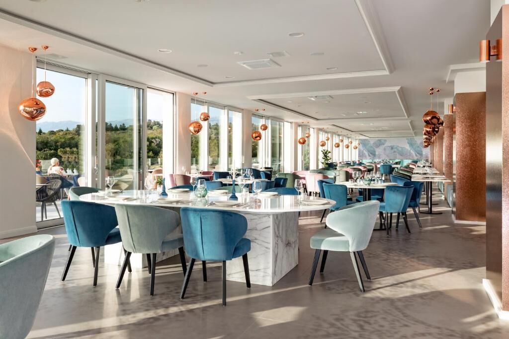 A modern dining area with round tables and blue chairs, large windows offering a view of the outdoors, and copper pendant lighting.