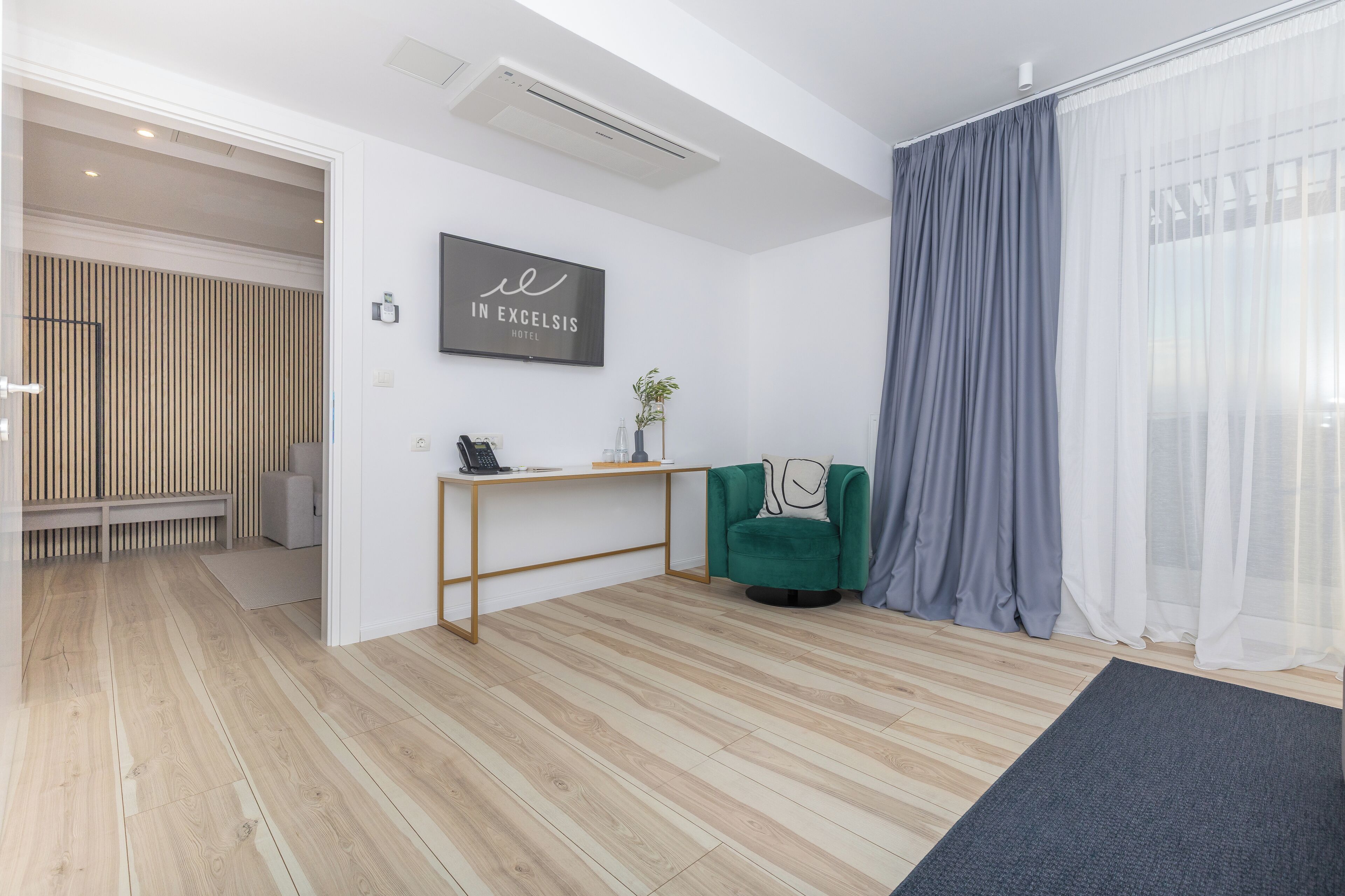 Una habitación de hotel moderna con suelo de madera, un sillón verde, un escritorio con una computadora portátil y un ventanal con cortinas translúcidas.