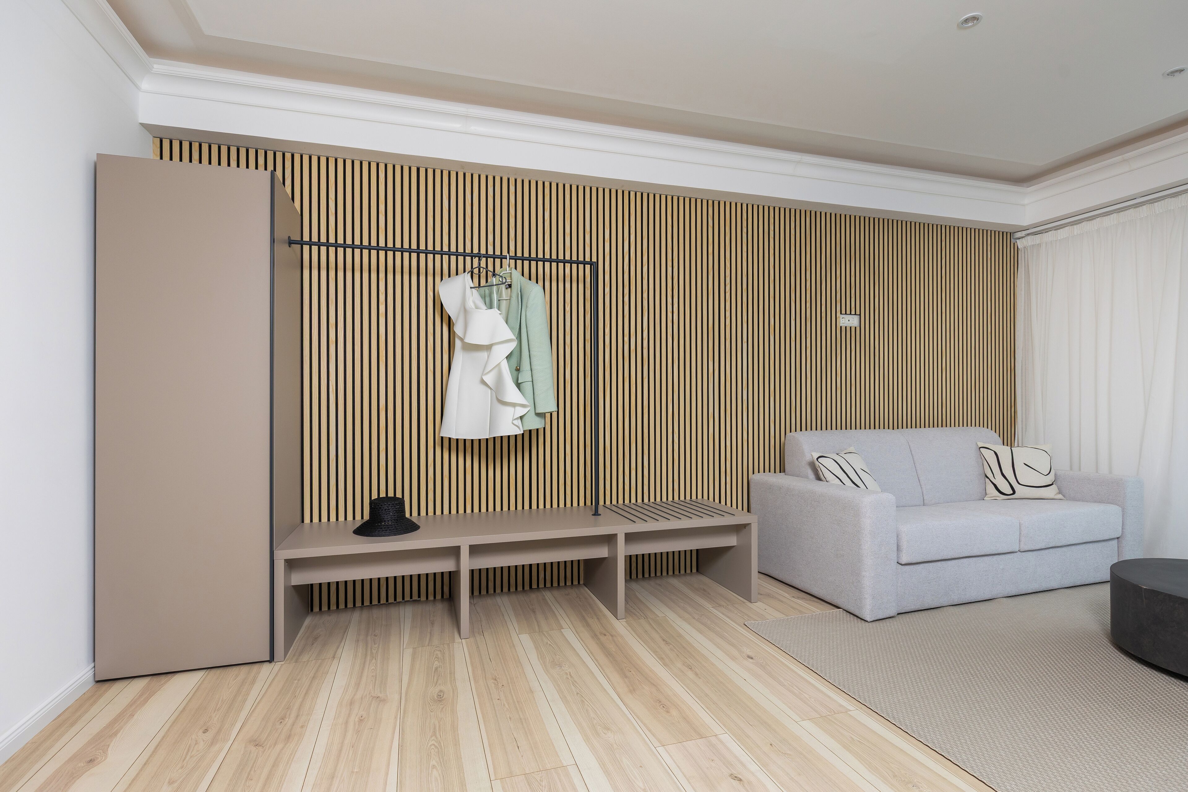 A modern living room with a grey sofa, a bench, and a clothing rack against a wooden slat wall.