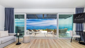 A modern hotel room with a wooden floor, a sofa, a small table, and a TV. A large sliding glass door leads to a balcony with a view of the sea and a lounge area with chairs and a table. - Hotel In Excelsis (Novalja)