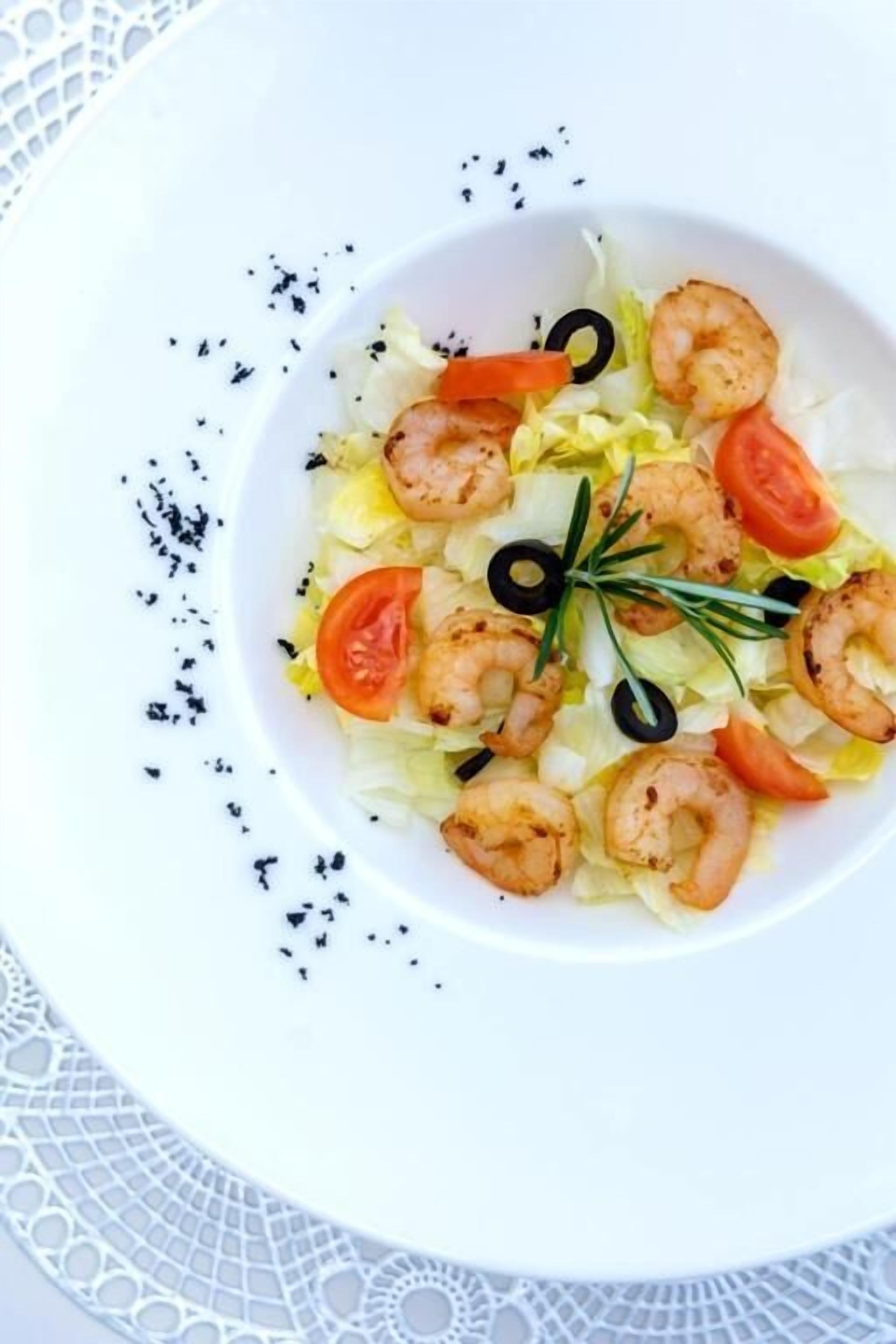 A white plate with a salad of lettuce, tomatoes, and shrimp, garnished with black olives and rosemary.