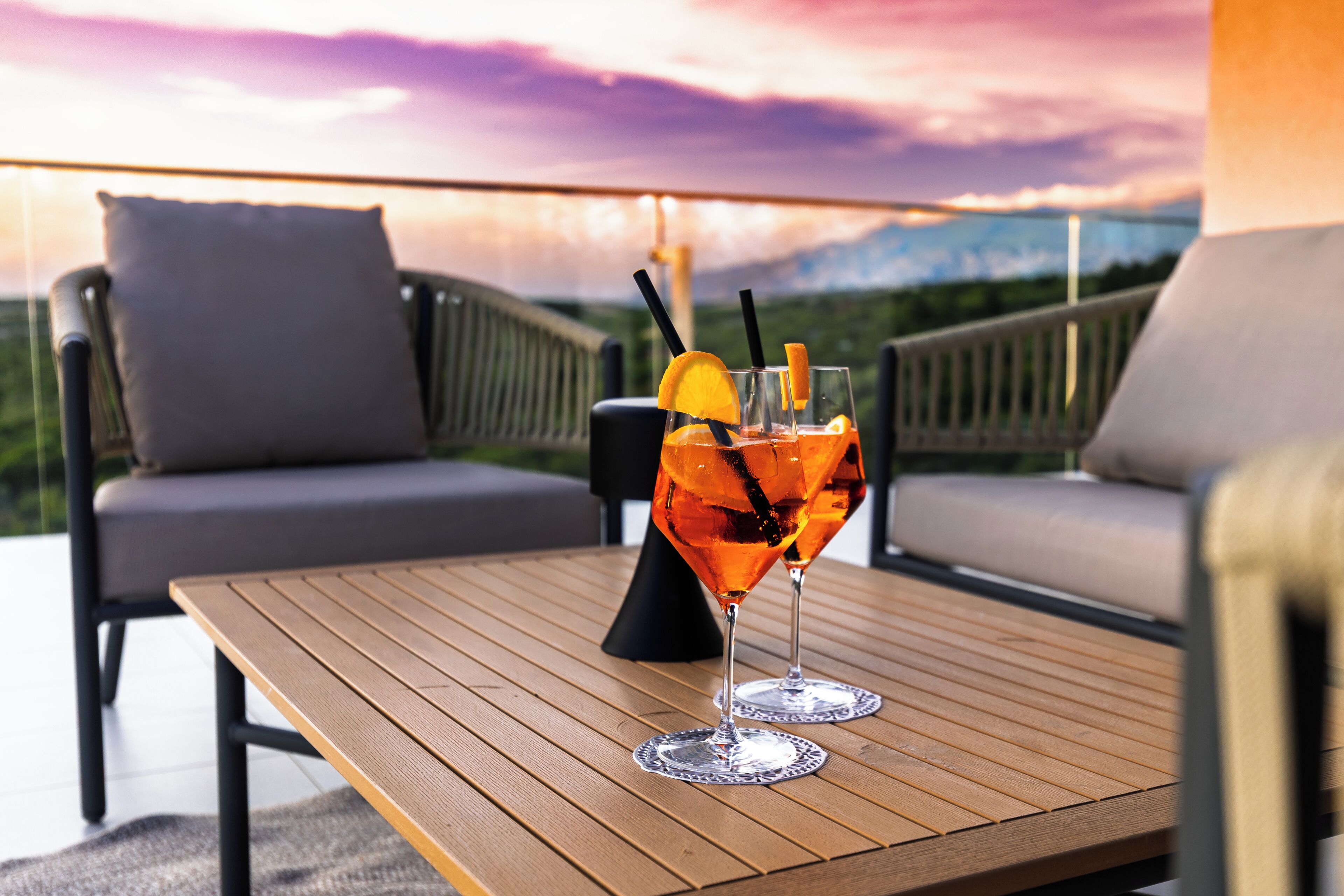 Una mesa de madera con dos vasos de jugo de naranja, un cojín y una silla en un balcón con vistas panorámicas.
