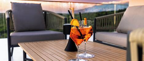 Una mesa de madera con dos vasos de jugo de naranja, un cojín y una silla en un balcón con vistas panorámicas.
