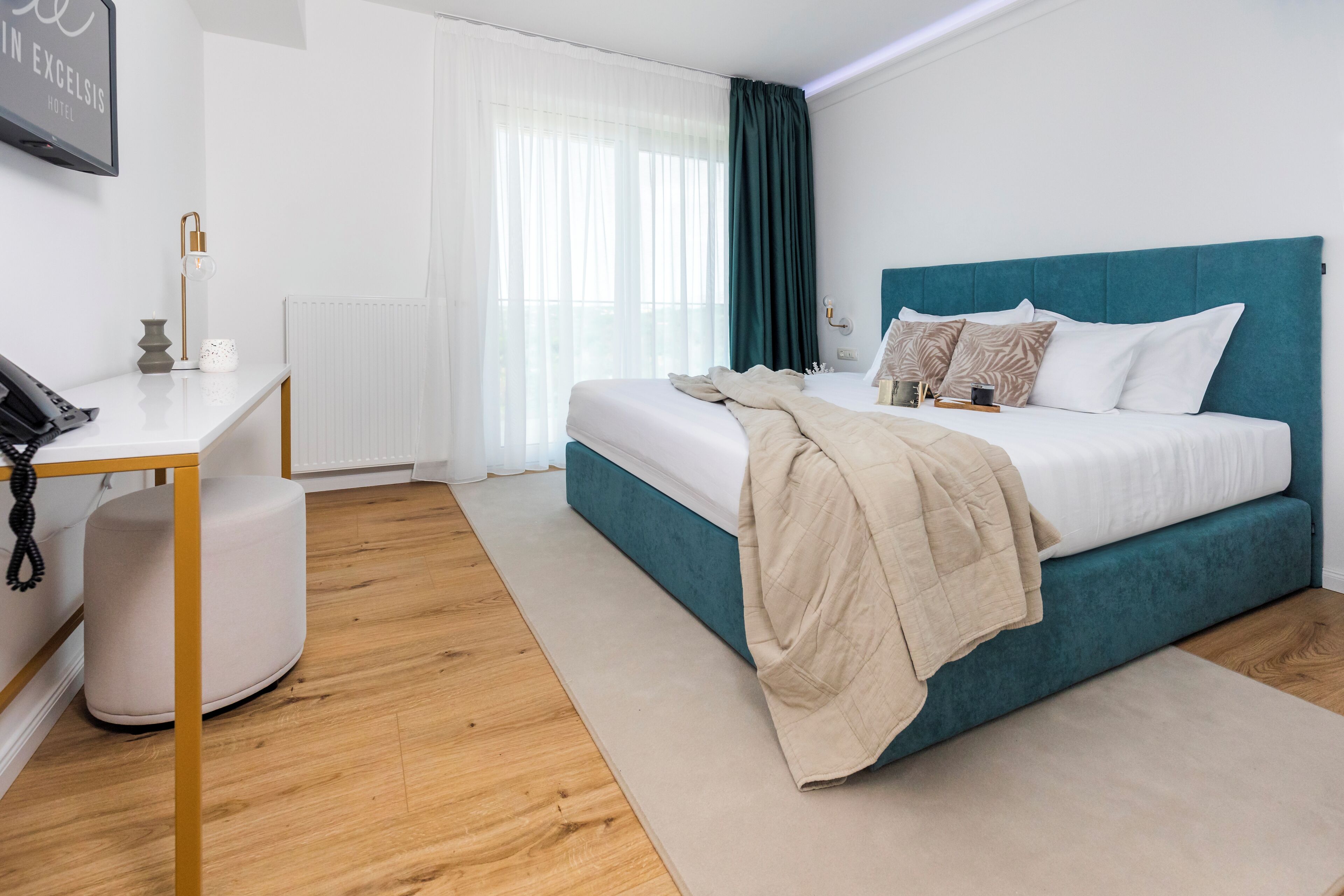 A modern hotel room with a teal bed, white walls, and a wooden floor.