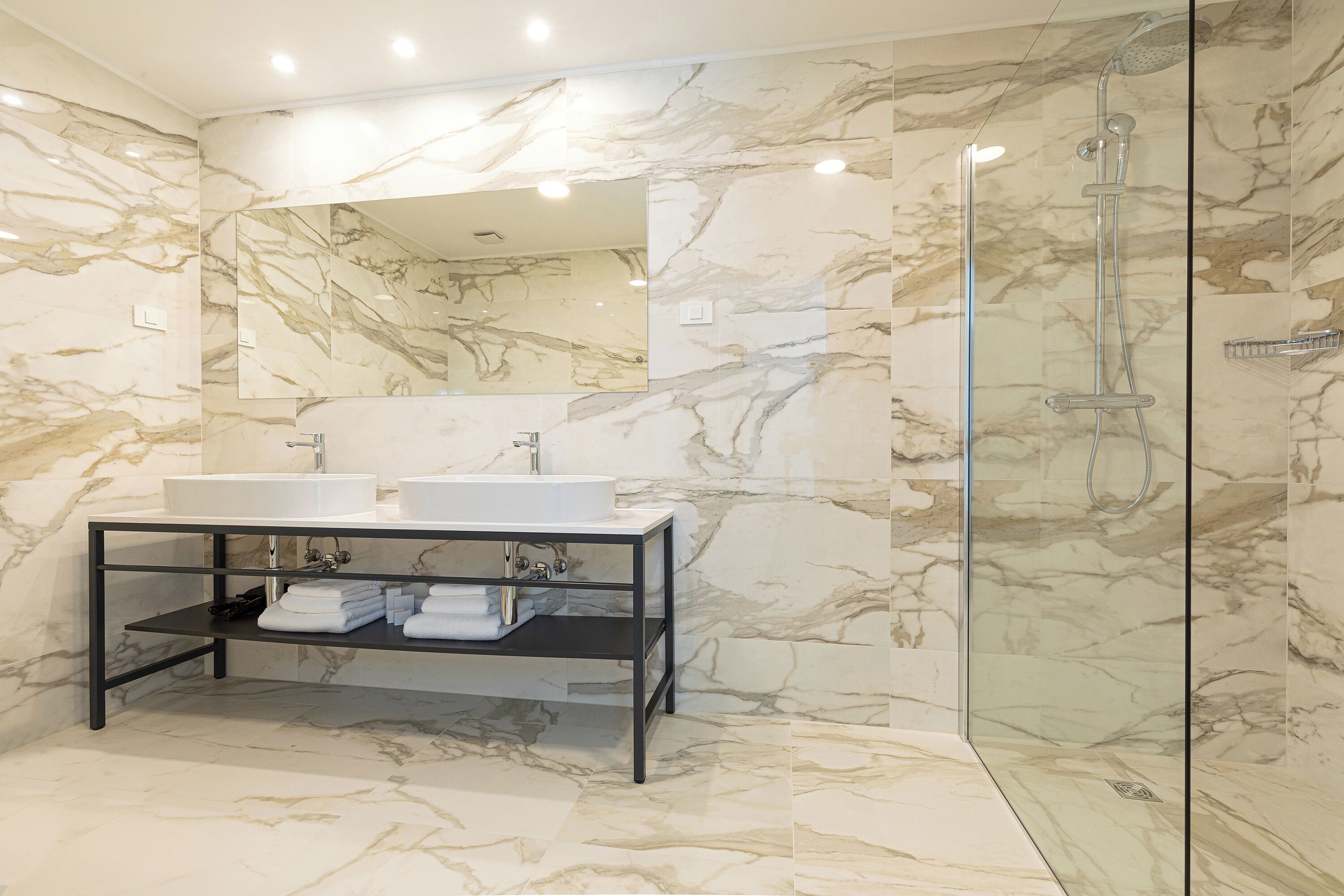 A modern bathroom with a marble wall, a large mirror, a double sink vanity, and a glass-enclosed shower.