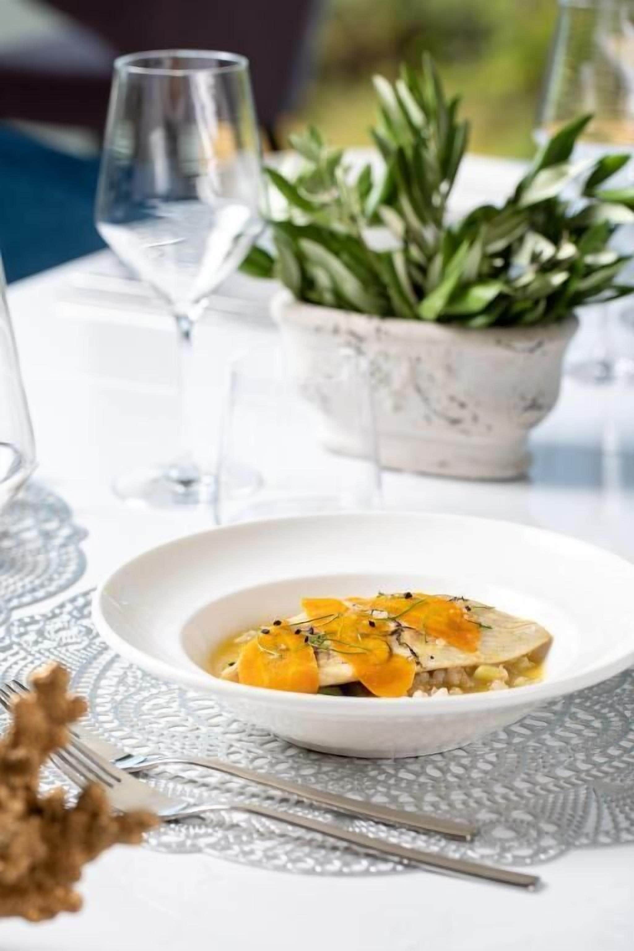 Une assiette blanche contenant de la nourriture, posée sur une table, entourée de verres à vin et d’une plante en pot.