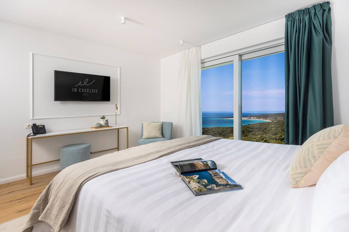 A modern hotel room with a large bed, a desk, and a TV displaying the hotel name.