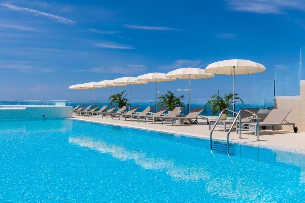 A swimming pool with sun loungers and umbrellas, surrounded by palm trees and a clear sky.