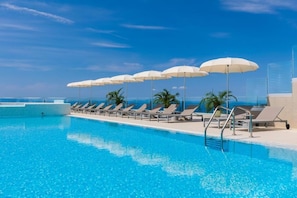 A swimming pool with sun loungers and umbrellas, surrounded by palm trees and a clear sky.