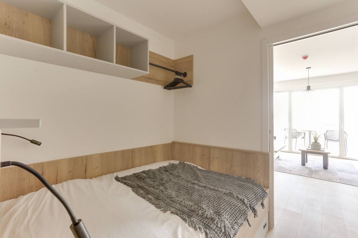 Ein Schlafzimmer mit einem Bett, einem Holz-Kopfbrett, einem Regal mit Kleiderhaken und Blick auf einen Balkon mit Stühlen und einem Tisch.