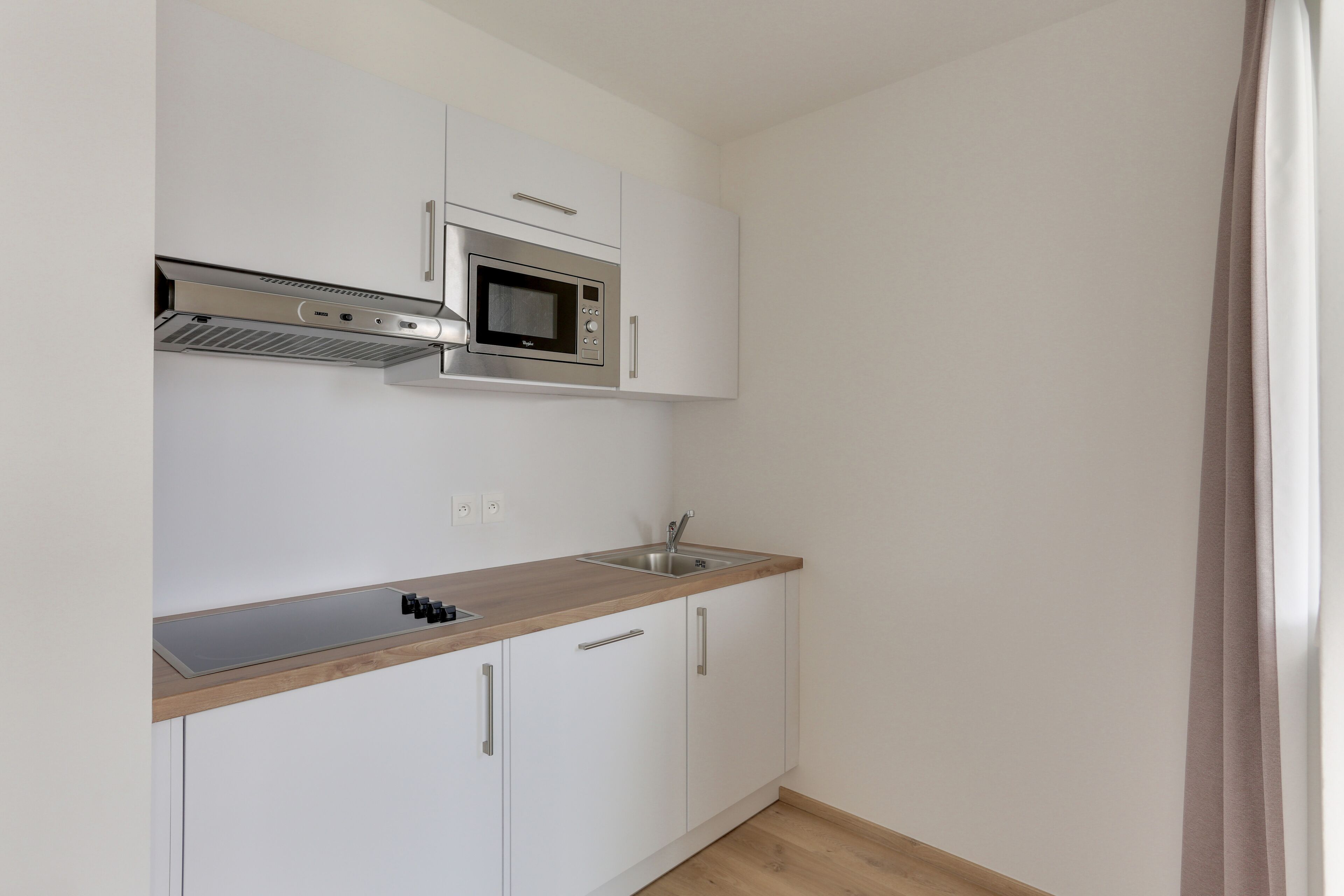 A compact kitchen with white cabinets, a stainless steel microwave, a built-in oven, and a wooden countertop.